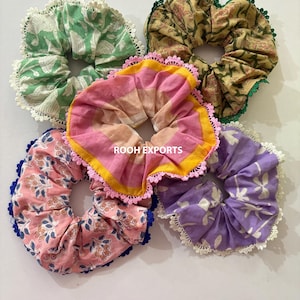 Op de afbeelding: Vijf kleurrijke scrunchies met bloemen- en patroonontwerpen. De scrunchies hebben verschillende kleuren, waaronder groen, roze, geel en paars, en zijn afgewerkt met kant. De scrunchies zijn gemaakt van stof en zijn ontworpen om haar vast te houden.
