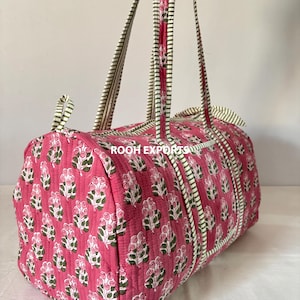 May include: A pink quilted duffel bag with a floral pattern. The bag features white and green striped handles and trim. The bag is cylindrical and has a zipper closure.