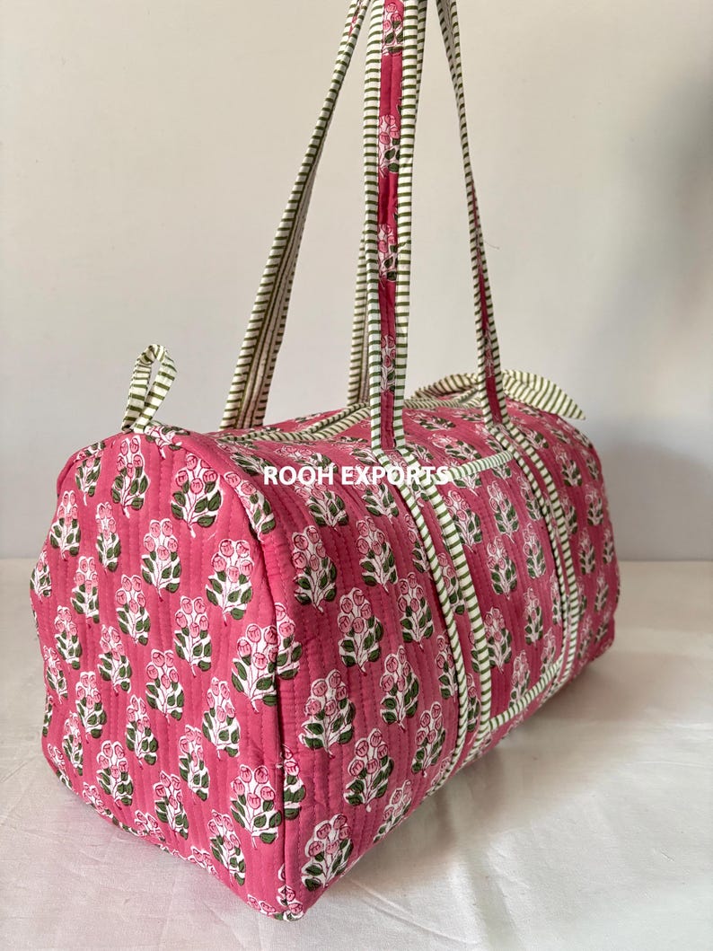 May include: A pink quilted duffel bag with a floral pattern. The bag features white and green striped handles and trim. The bag is cylindrical and has a zipper closure.