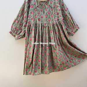 Puede incluir: Un vestido de estampado floral verde y rosa con un lazo en el escote y mangas abullonadas. El vestido est&aacute; colgado en una percha de madera.