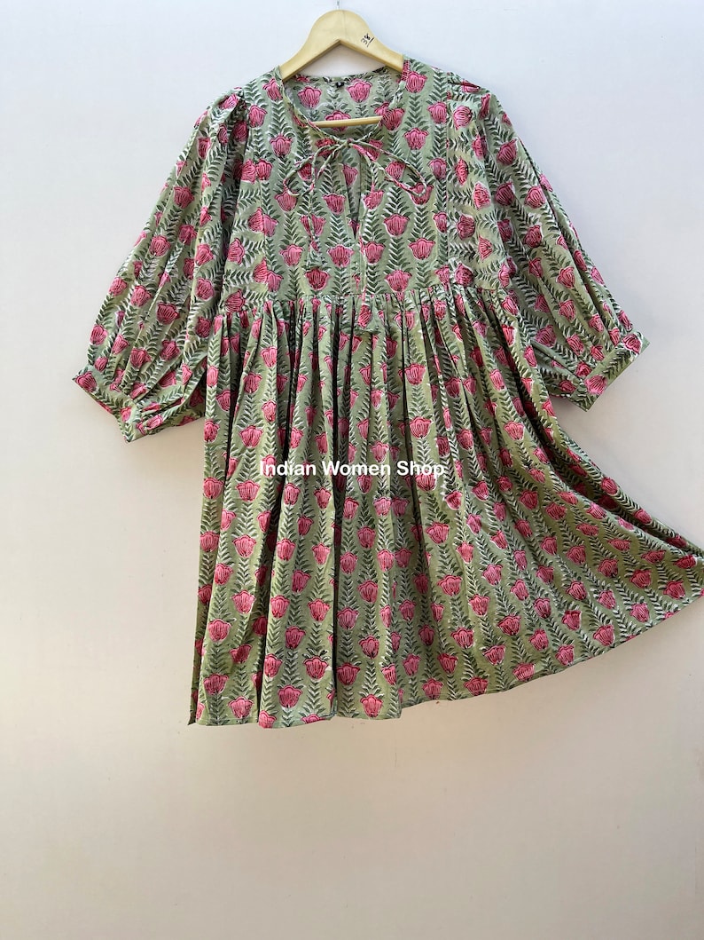 Puede incluir: Un vestido de estampado floral verde y rosa con un lazo en el escote y mangas abullonadas. El vestido est&aacute; colgado en una percha de madera.