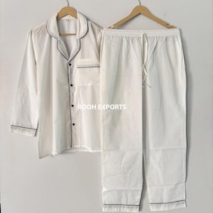 Peut inclure: Ensemble de pyjama blanc avec bordure noire, comprenant une chemise boutonnée et un pantalon à cordon. La chemise a une poche poitrine et le texte "ROOH EXPORTS". L'ensemble est présenté sur des cintres en bois.