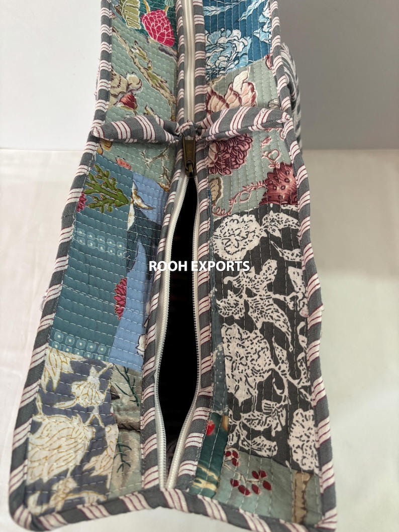 May include: Close-up of a quilted tote bag with a patchwork design. The bag features a variety of floral patterns in shades of blue, gray, and pink. A white zipper runs down the center, and the edges are trimmed with a striped fabric. The text "ROOH EXPORTS" is visible.