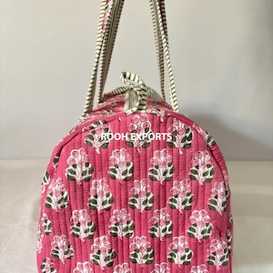 May include: A pink quilted travel bag with a floral pattern. The bag features a repeating design of white and pink flowers with green leaves. The handles are striped in white, pink, and green. The bag has a zipper closure.