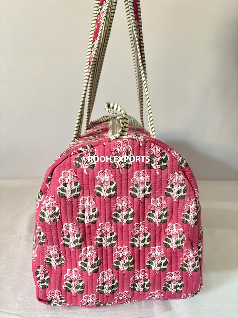 May include: A pink quilted travel bag with a floral pattern. The bag features a repeating design of white and pink flowers with green leaves. The handles are striped in white, pink, and green. The bag has a zipper closure.