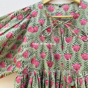Puede incluir: Un vestido de estampado floral verde y rosa con un lazo en el escote. El vestido tiene una falda fruncida y mangas 3/4.