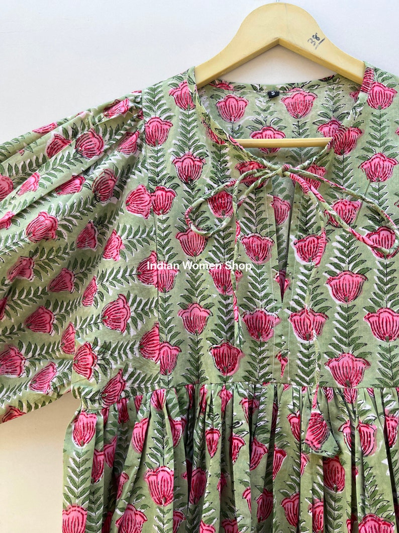 Puede incluir: Un vestido de estampado floral verde y rosa con un lazo en el escote. El vestido tiene una falda fruncida y mangas 3/4.