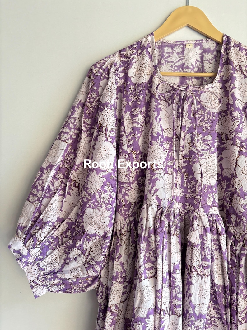 May include: A purple and white floral print cotton dress with a gathered skirt and puffed sleeves. The dress has a tie at the neckline.