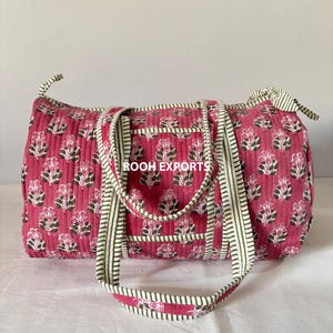 May include: A quilted duffel bag in a vibrant pink hue, adorned with a floral pattern in white and green. The bag features sturdy handles and trim with a green and white striped design. The text "ROOH EXPORTS" is visible.