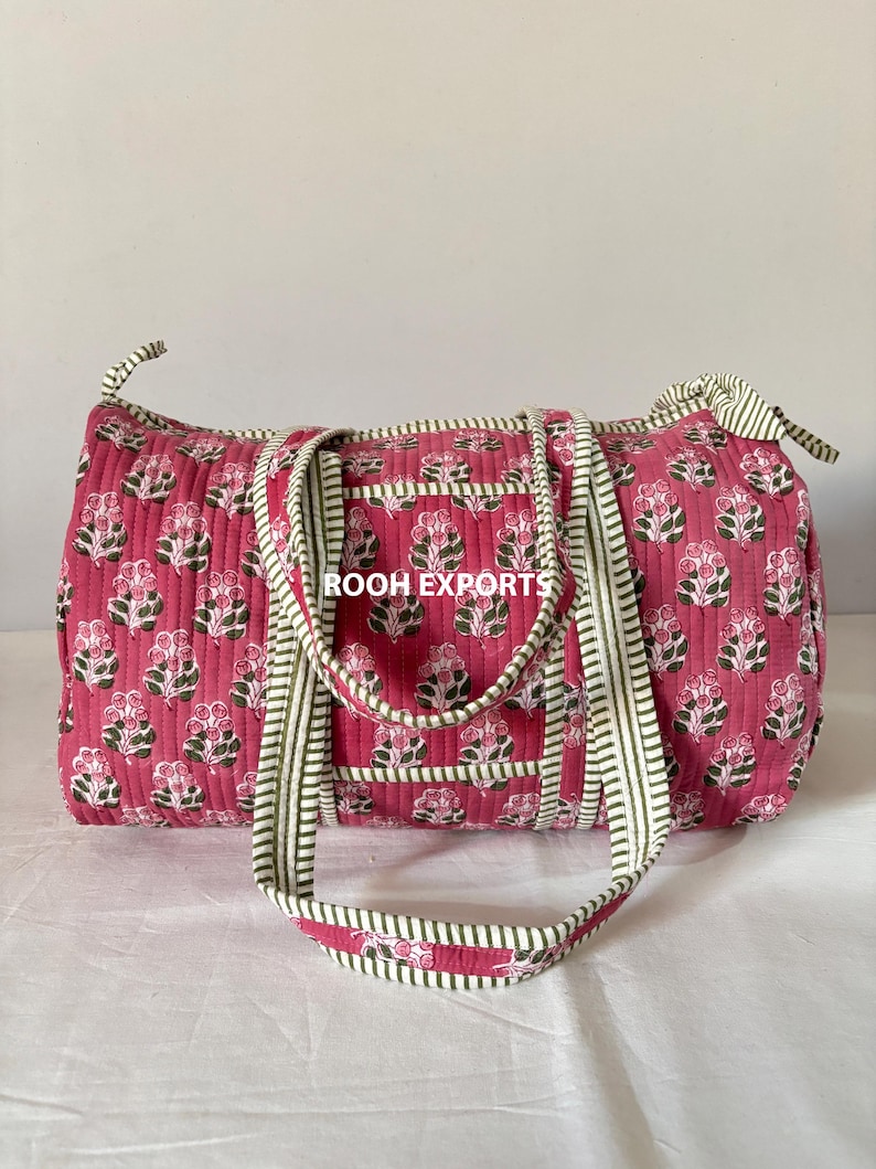 May include: A quilted duffel bag in a vibrant pink hue, adorned with a floral pattern in white and green. The bag features sturdy handles and trim with a green and white striped design. The text "ROOH EXPORTS" is visible.