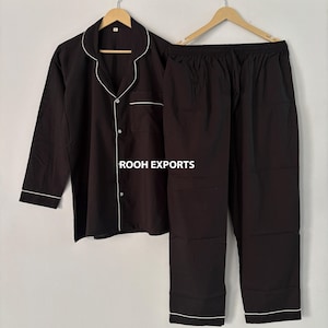 Puede incluir: Conjunto de pijama negro con ribete blanco. La camisa abotonada de manga larga y el pantalón a juego se muestran en perchas de madera. El texto "ROOH EXPORTS" está impreso en la camisa.