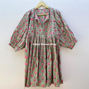 Puede incluir: Un vestido de estampado floral verde y rosa con cuello en V y mangas 3/4. El vestido tiene una cintura fruncida y una falda fluida. El vestido est&aacute; colgado en una percha de madera. "Indian Women Shop" est&aacute; impreso en el vestido.