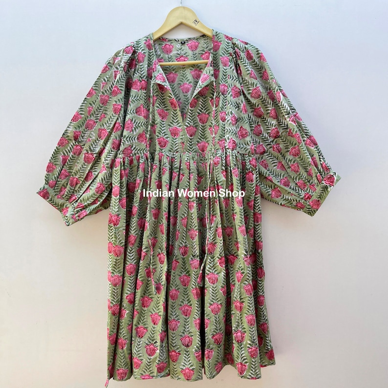 Puede incluir: Un vestido de estampado floral verde y rosa con cuello en V y mangas 3/4. El vestido tiene una cintura fruncida y una falda fluida. El vestido est&aacute; colgado en una percha de madera. "Indian Women Shop" est&aacute; impreso en el vestido.