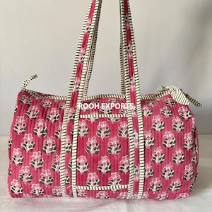 May include: A quilted duffel bag in a vibrant pink hue, adorned with a repeating floral pattern in white and green. The bag features matching pink handles and trim with a striped design. The bag is designed for travel or everyday use.