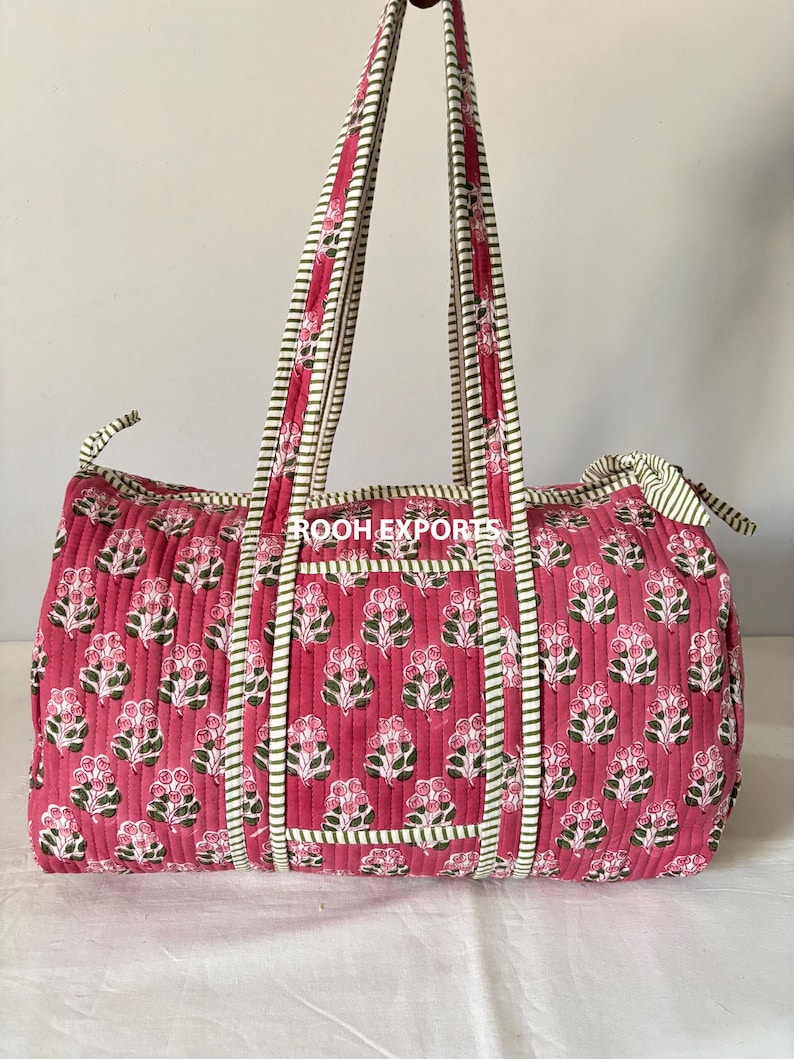 May include: A quilted duffel bag in a vibrant pink hue, adorned with a repeating floral pattern in white and green. The bag features matching pink handles and trim with a striped design. The bag is designed for travel or everyday use.
