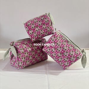 May include: Three quilted cosmetic bags. The bags are stacked and feature a magenta background with a white and olive green leaf pattern. Each bag has a striped zipper and matching striped fabric ties.