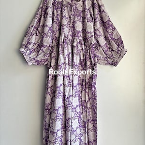 May include: A long, purple and white floral print dress with a gathered waist and long, puffed sleeves. The dress has a tie at the waist and is made of a lightweight fabric.