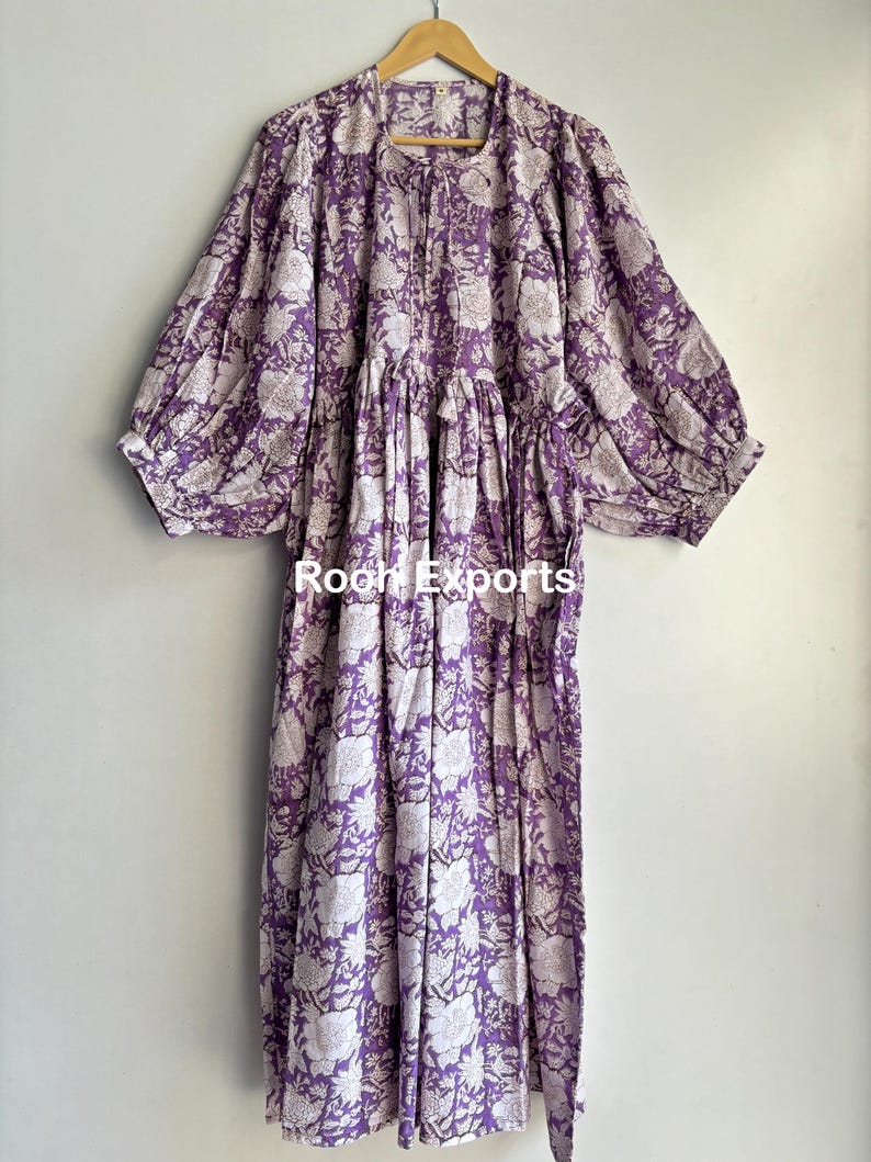 May include: A long, purple and white floral print dress with a gathered waist and long, puffed sleeves. The dress has a tie at the waist and is made of a lightweight fabric.