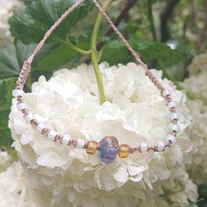 Puede incluir: Una pulsera hecha a mano con un cordón beige y cuentas blancas. La pulsera presenta una cuenta central azul y marrón, flanqueada por dos cuentas de color ámbar. La pulsera se exhibe sobre flores blancas.