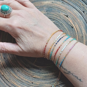 Può includere: Una mano che indossa una varietà di braccialetti colorati con perline e dettagli dorati. I braccialetti sono legati con stringhe e hanno perline di diversi colori, tra cui rosso, arancione, giallo, verde, blu e bianco.