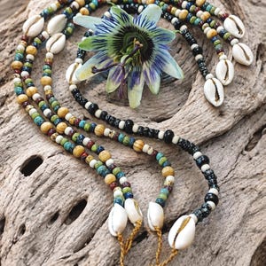 Può includere: Una collana a più fili con piccole perline rotonde nei toni del blu, verde, giallo e nero, intervallate da conchiglie di ciprea bianche. La collana è disposta in un motivo circolare attorno a un singolo fiore di passiflora blu e bianco.