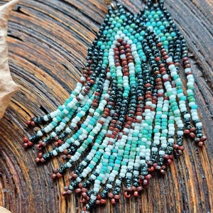 Op de afbeelding: Een paar lange, geperde oorbellen met een turquoise, wit, zwart en bruin kleurenschema. De oorbellen zijn gemaakt van kleine, ronde kralen en hebben een franje-achtig ontwerp.