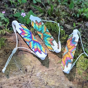 Può includere: Tre braccialetti in macramè colorati con intricati motivi geometrici. Ogni braccialetto presenta una base bianca con vibranti tonalità arcobaleno intrecciate. I braccialetti sono esposti su una superficie di legno con uno sfondo naturale all'aperto.