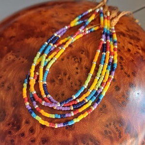 Peut inclure: Plusieurs colliers de perles colorées. Chaque collier présente un arc-en-ciel de couleurs, notamment rouge, orange, jaune, vert, bleu et violet. Les perles sont enfilées avec des accents dorés. Les colliers sont présentés sur une surface en bois.