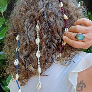 Puede incluir: Primer plano de cabello castaño rizado adornado con tres hebras de accesorios para el cabello de conchas. Una hebra es azul, otra blanca y otra burdeos. Cada hebra presenta múltiples conchas blancas. El fondo es una planta verde frondosa.