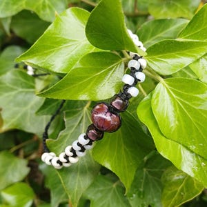 Peut inclure: Un bracelet fait à la main composé de perles blanches et noires alternées, avec des perles rouges foncées plus grandes. Le bracelet est monté sur un cordon noir. Le fond est un feuillage vert luxuriant.
