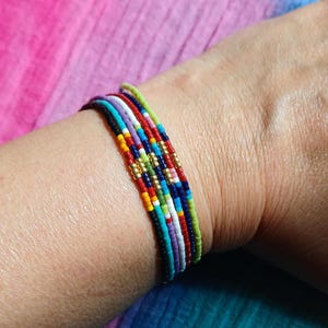 Peut inclure: Plusieurs bracelets de perles colorées. Les bracelets présentent une variété de couleurs, notamment le rouge, le bleu, le vert, le jaune et le blanc, avec des perles dorées parsemées. Les bracelets sont empilés les uns sur les autres.