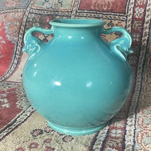 Könnte beinhalten: Eine tealfarbene Keramikvase mit elefantförmigen Griffen. Die Vase hat einen runden Körper und eine glatte, glänzende Oberfläche.
