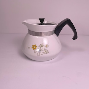 Puede incluir: Cafetera de cerámica blanca con asa y tapa negras. La cafetera presenta una banda plateada y diseños florales en amarillo y naranja. El pico está en el lado izquierdo.