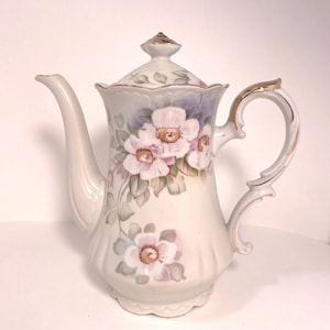 May include: A white porcelain teapot with a lid, featuring hand-painted pink and purple floral designs. The teapot has a curved spout, a gold-trimmed handle, and a scalloped base. A decorative piece for serving tea.