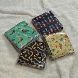 May include: Four stacks of square paper napkins in clear plastic packaging. The napkins feature various patterns: roller skates, fireworks, candy corn worms, and woodland animals. The napkins are on a white, textured surface.