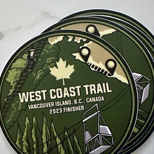 May include: A green and white round sticker with the text "West Coast Trail" and "Vancouver Island, B.C., Canada 2023 Finisher". The sticker features a stylized illustration of a mountain with a cable car and trees.