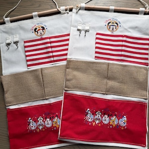 May include: Two white fabric organizers with wooden dowels and rope hangers. Each has red and white striped pockets, burlap pockets, and red pockets with embroidered Disney characters. The top features the "Disney Cruise Line" logo.