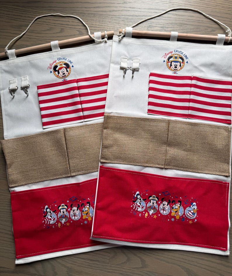 May include: Two white fabric organizers with wooden dowels and rope hangers. Each has red and white striped pockets, burlap pockets, and red pockets with embroidered Disney characters. The top features the "Disney Cruise Line" logo.