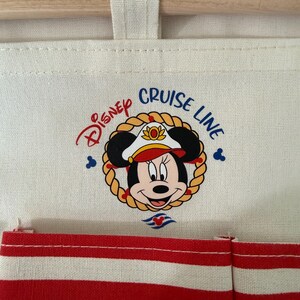 May include: Cream-colored fabric organizer with a wooden rod and red and white striped pockets. The Disney Cruise Line logo features Minnie Mouse in a captain's hat, surrounded by a rope border.