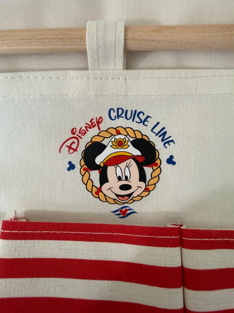 May include: Cream-colored fabric organizer with a wooden rod and red and white striped pockets. The Disney Cruise Line logo features Minnie Mouse in a captain's hat, surrounded by a rope border.