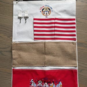 May include: A white fabric organizer with red and white striped pockets and a red bottom pocket featuring Disney characters. The top has a Disney Cruise Line logo and two metal hooks. A wooden rod and rope are at the top.