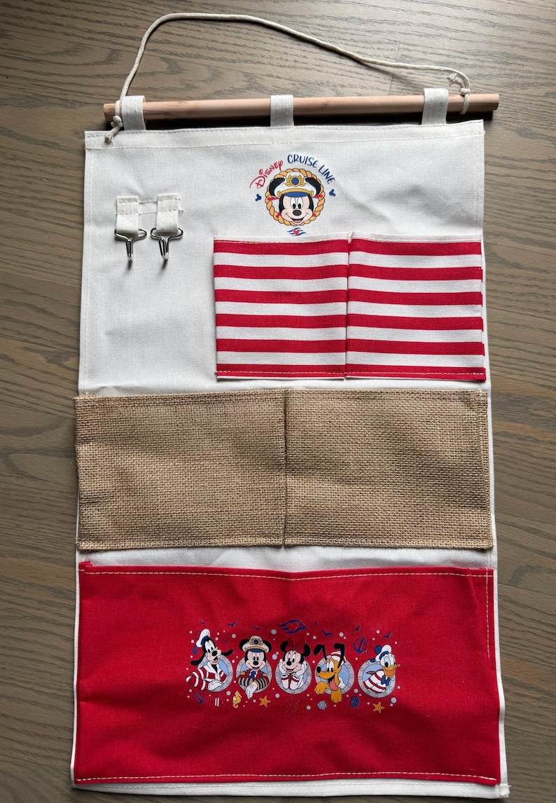 May include: A white fabric organizer with red and white striped pockets and a red bottom pocket featuring Disney characters. The top has a Disney Cruise Line logo and two metal hooks. A wooden rod and rope are at the top.