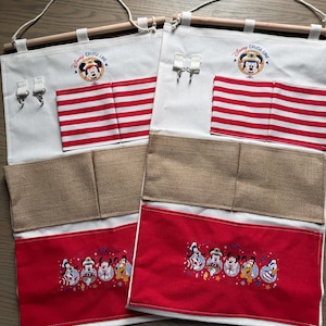 May include: Two white fabric organizers with wooden dowels and hanging loops. Each has red and white striped pockets, a burlap pocket, and a red pocket with embroidered Disney characters. The top features a Mickey Mouse logo and the words "Disney Cruise Line".