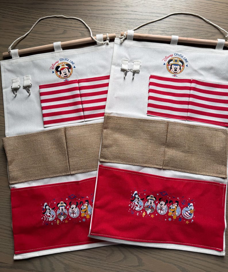 May include: Two white fabric organizers with wooden dowels and hanging loops. Each has red and white striped pockets, a burlap pocket, and a red pocket with embroidered Disney characters. The top features a Mickey Mouse logo and the words "Disney Cruise Line".