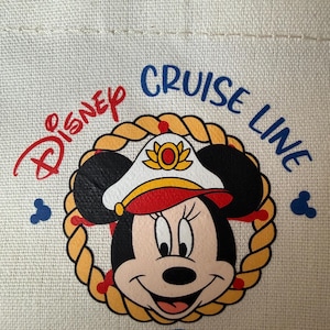 May include: Cream-colored tote bag featuring a Minnie Mouse graphic in a captain's hat, surrounded by a rope design. The words "Disney Cruise Line" are printed above the graphic in red and blue. Two Mickey Mouse silhouettes are on the sides.
