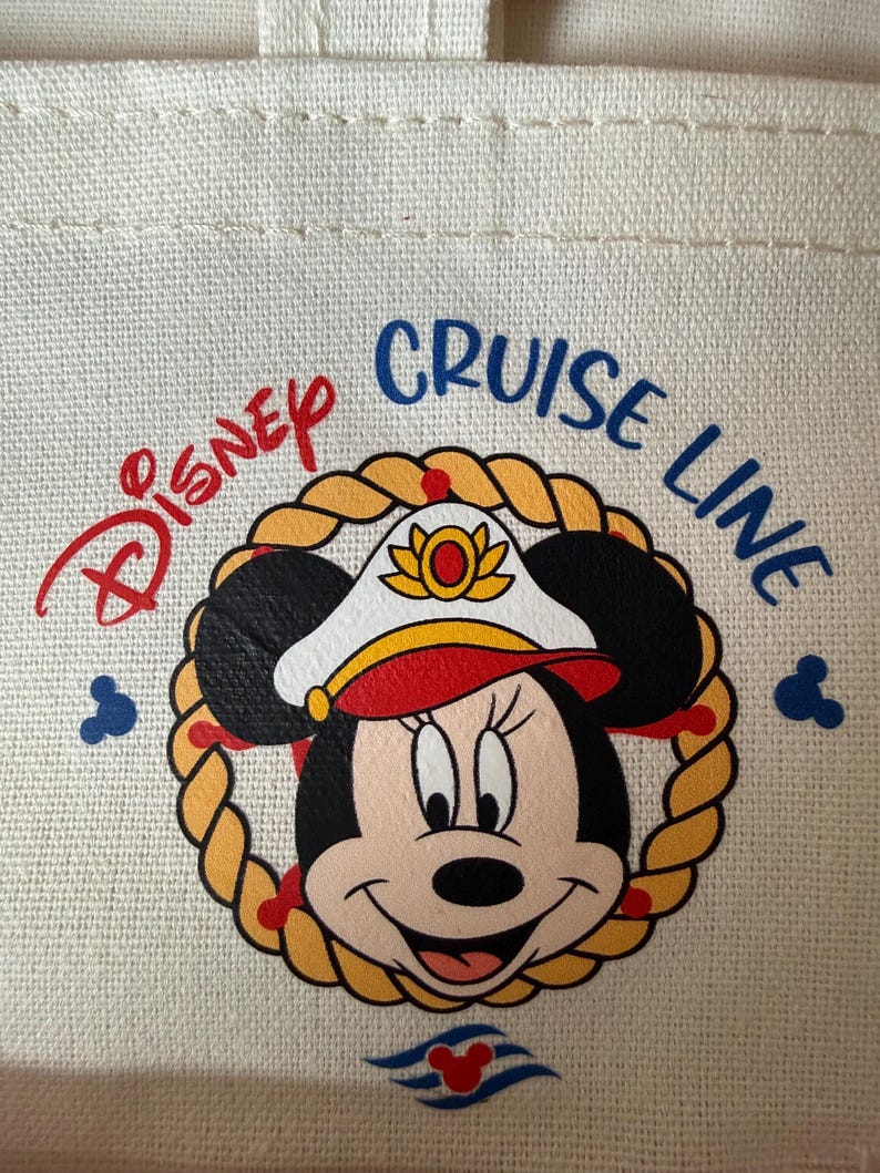 May include: Cream-colored tote bag featuring a Minnie Mouse graphic in a captain's hat, surrounded by a rope design. The words "Disney Cruise Line" are printed above the graphic in red and blue. Two Mickey Mouse silhouettes are on the sides.