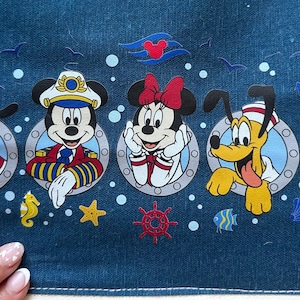 May include: A denim fabric featuring cartoon characters Goofy, Mickey Mouse, Minnie Mouse, Pluto, and Donald Duck in nautical-themed designs. The characters are framed in portholes with nautical accents like anchors and starfish.