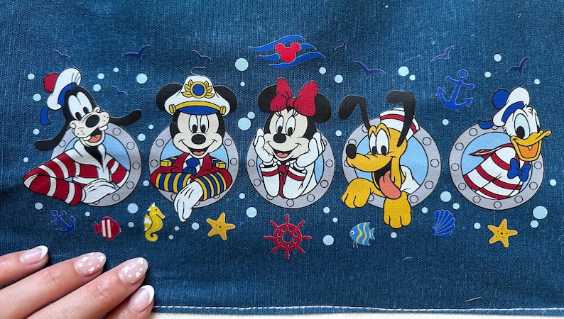 May include: A denim fabric featuring cartoon characters Goofy, Mickey Mouse, Minnie Mouse, Pluto, and Donald Duck in nautical-themed designs. The characters are framed in portholes with nautical accents like anchors and starfish.