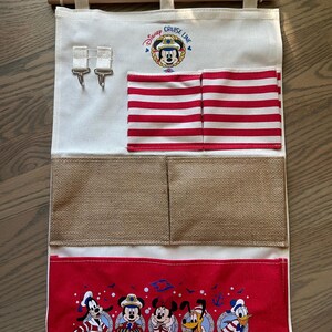 May include: A canvas wall organizer featuring the Disney Cruise Line logo and Mickey Mouse. It has red and white striped pockets, burlap pockets, and a red bottom panel with Disney characters. Includes a wooden dowel and hanging cord.