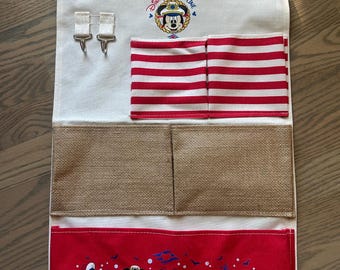 May include: A canvas wall organizer featuring the Disney Cruise Line logo and Mickey Mouse. It has red and white striped pockets, burlap pockets, and a red bottom panel with Disney characters. Includes a wooden dowel and hanging cord.